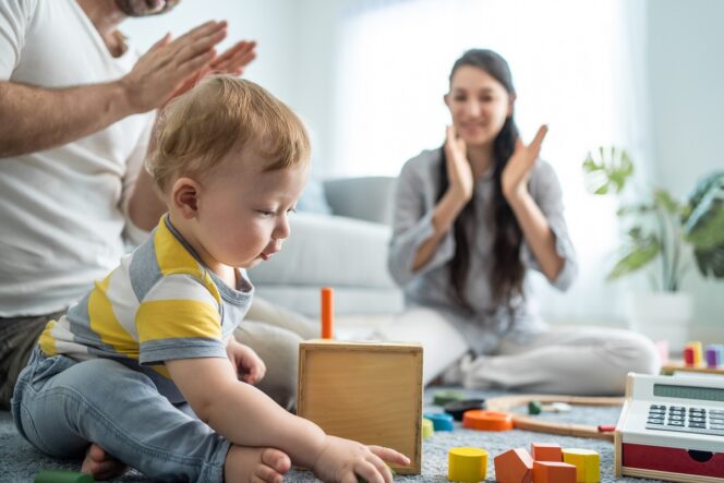 
 Caucasian loving parent looking at baby toddler playing in living room. Attractive couple mother and father watch young little infant son child's development. Activity relationship at home in house.